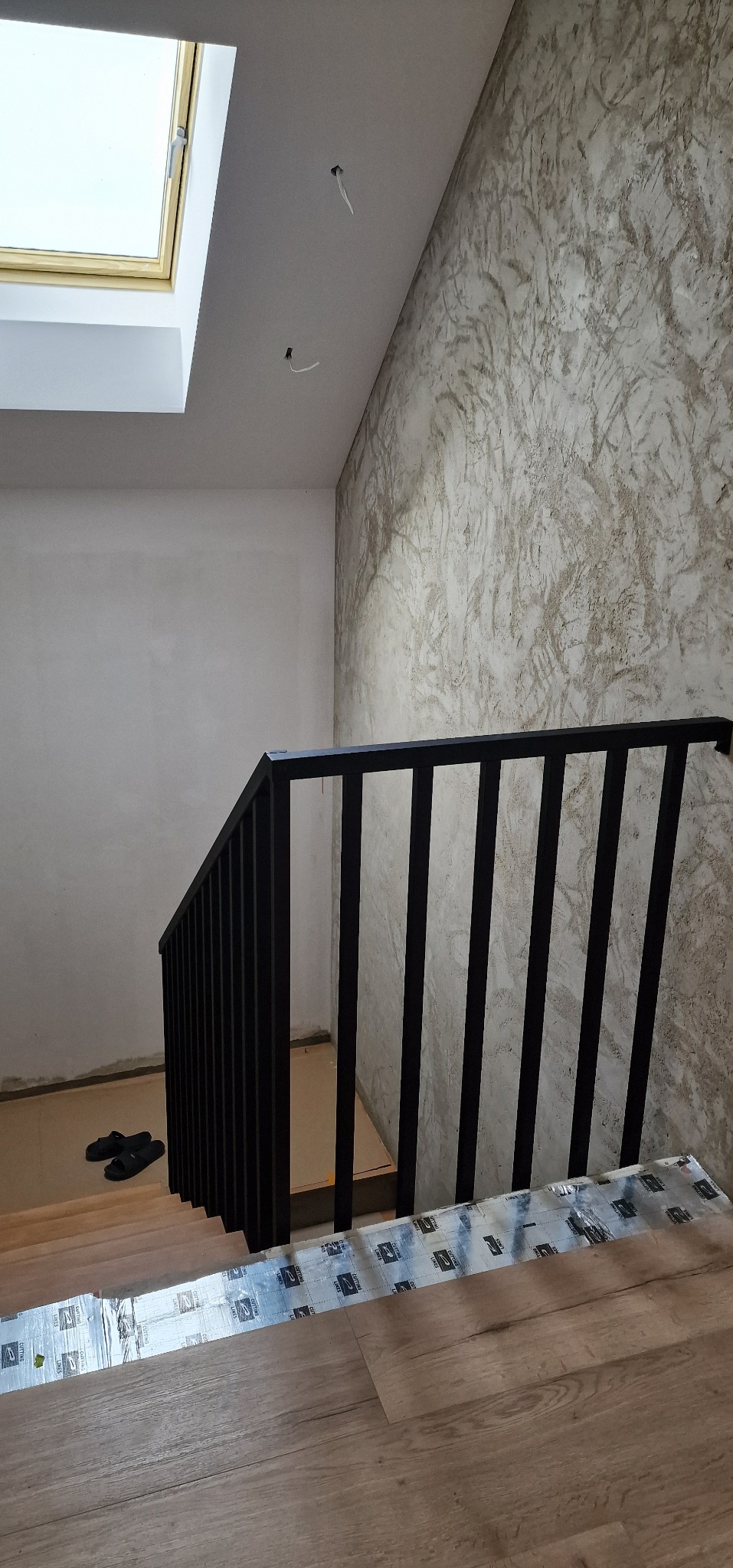 Nowoczesna, czarna balustrada schodowa z pionowymi szczeblami. Widoczne schody z drewnopodobnych paneli i dekoracyjna faktura ściany. Ujęcie z dołu, z oknem dachowym.