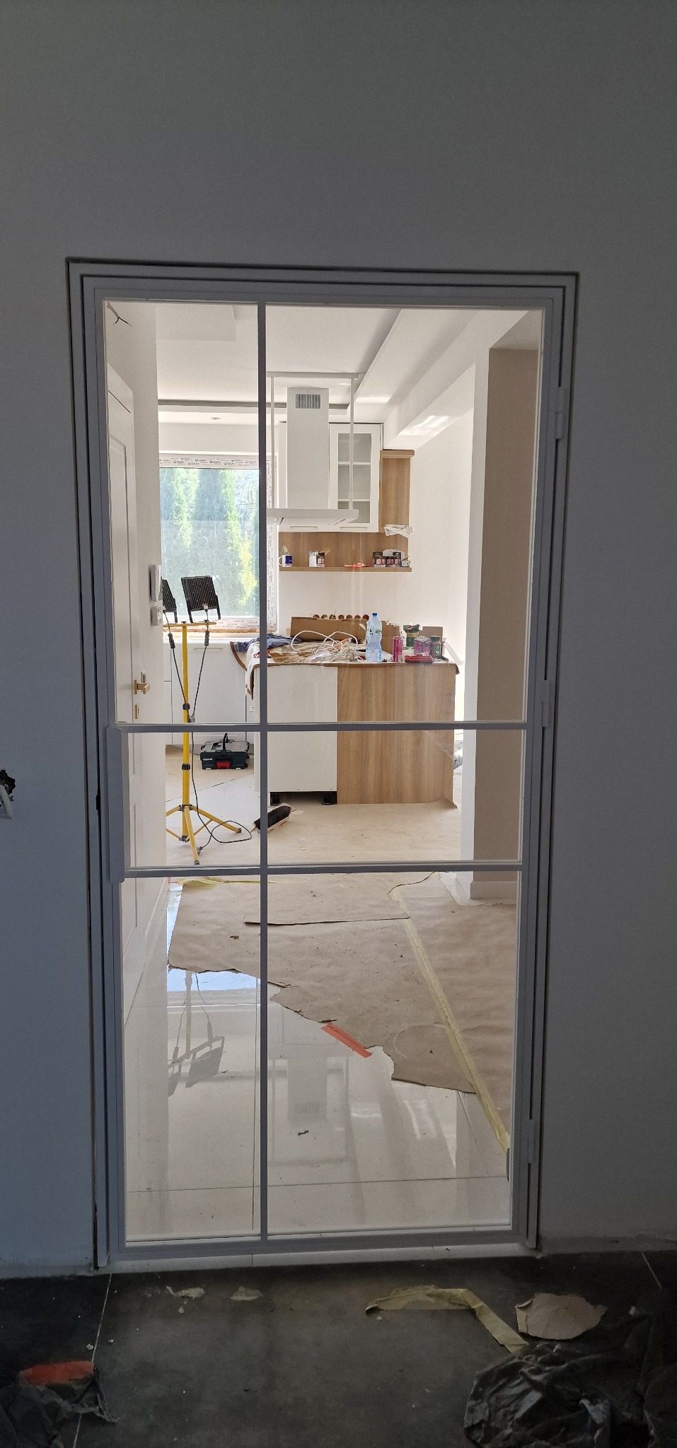 Nowoczesna, biała rama okienna z podziałem na cztery pola, widok na wnętrze kuchni w trakcie remontu. Oświetlenie budowlane, zabezpieczona podłoga.