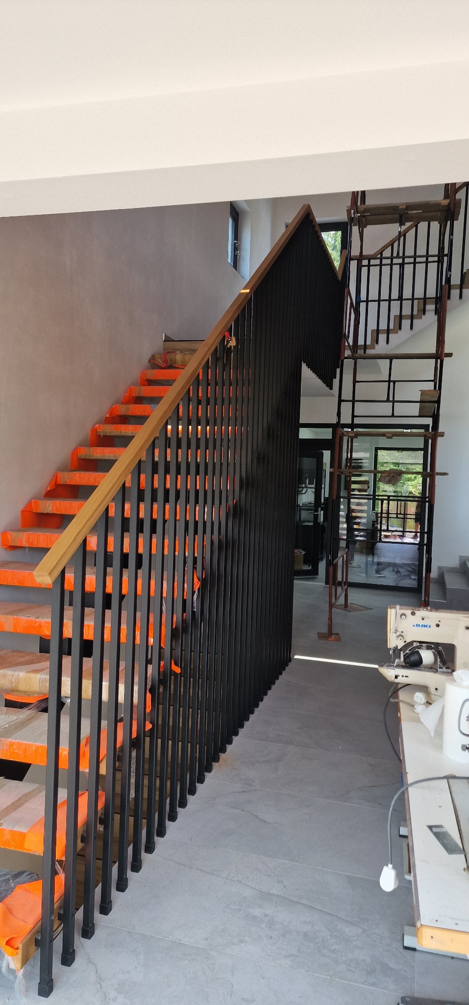 Nowoczesne schody z czarną balustradą i drewnianym poręczą. Stopnie zabezpieczone folią ochronną. W tle widoczne rusztowanie i maszyna do szycia.