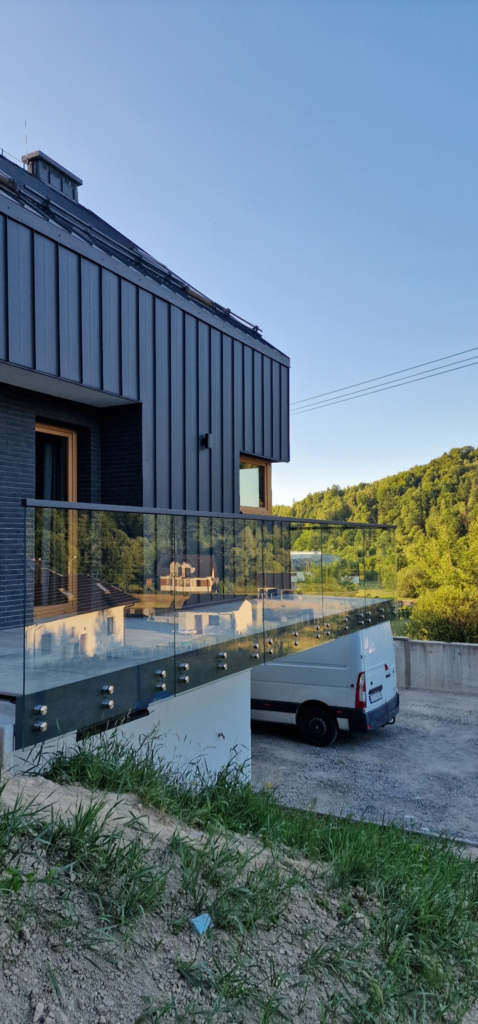 Szklana balustrada z mocowaniami punktowymi na schodach zewnętrznych nowoczesnego domu z blachy trapezowej, z widocznym samochodem dostawczym w tle.