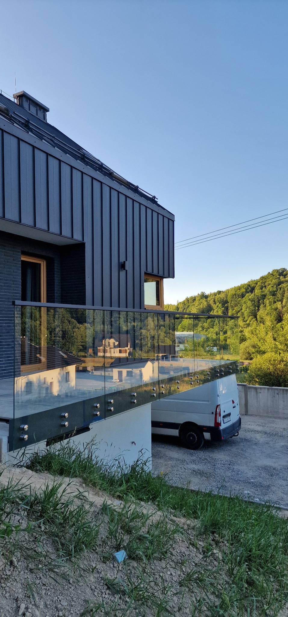 Nowoczesna balustrada szklana z mocowaniami punktowymi przy schodach zewnętrznych domu z grafitową elewacją. W tle zaparkowany biały bus.