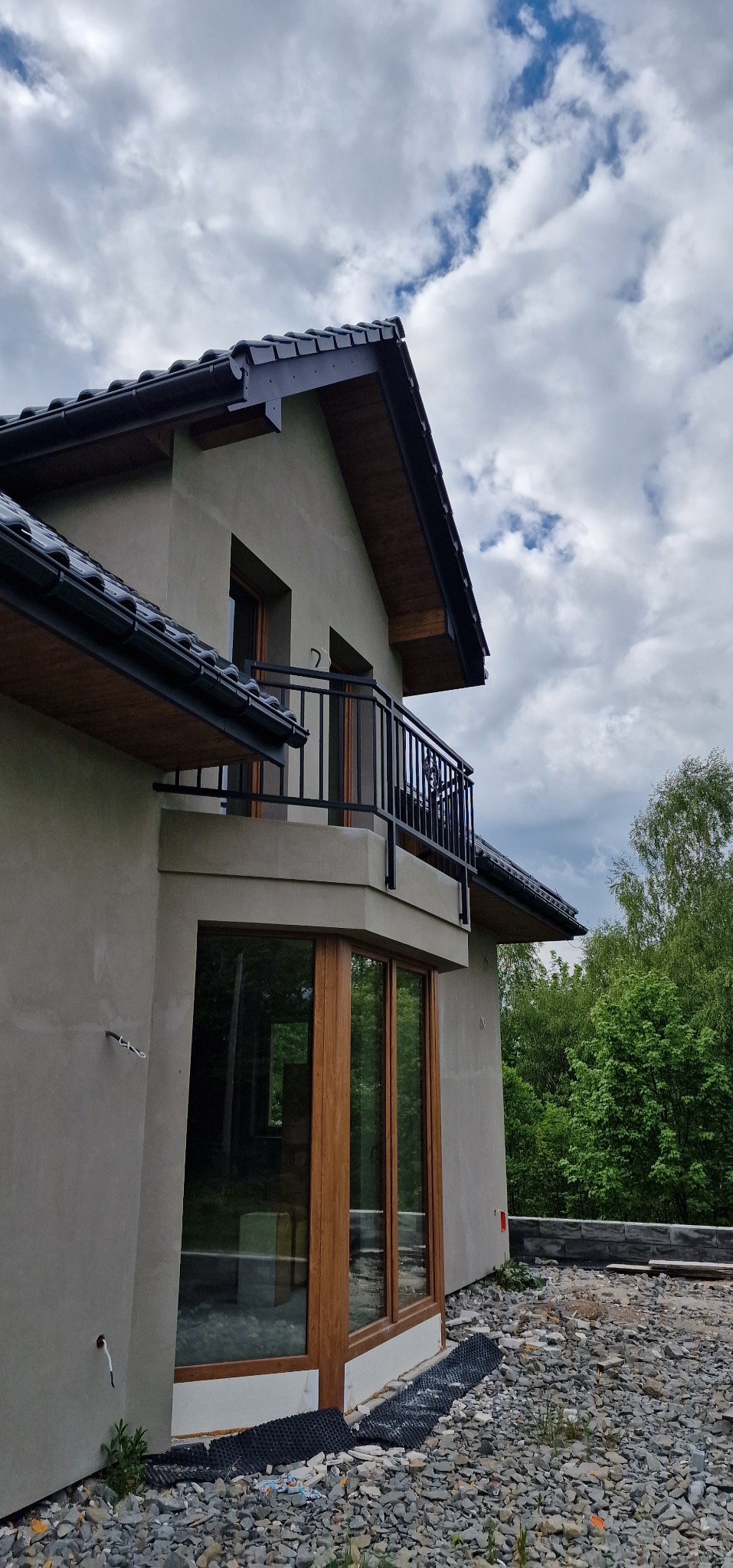Dom z balkonem z czarną, metalową balustradą na tle pochmurnego nieba. Widoczne drewniane okna i fragment dachu. Teren wokół domu w trakcie prac wykończeniowych.