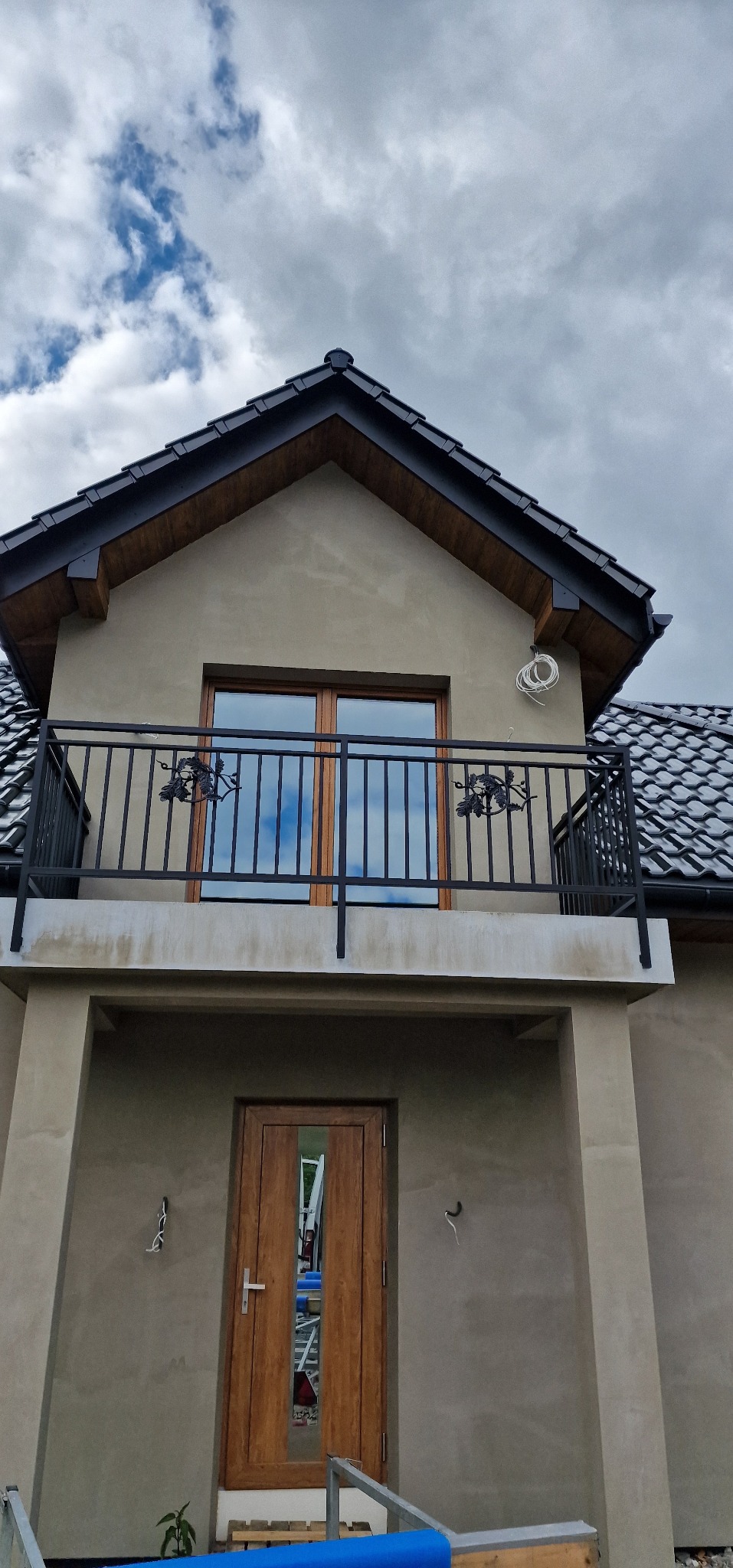 Dom z balkonem i czarną, metalową balustradą z motywem liści winogron. Widoczne drewniane okna i drzwi wejściowe. Budynek w trakcie wykończenia.