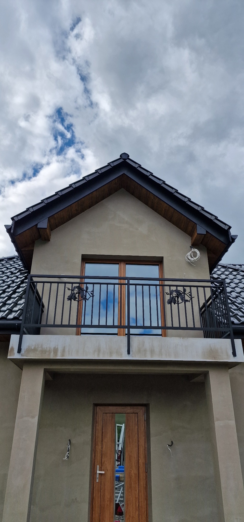 Dom z balkonem zabezpieczonym czarną, metalową balustradą z dekoracyjnymi elementami roślinnymi. W tle błękitne niebo z chmurami odbijające się w szybach.