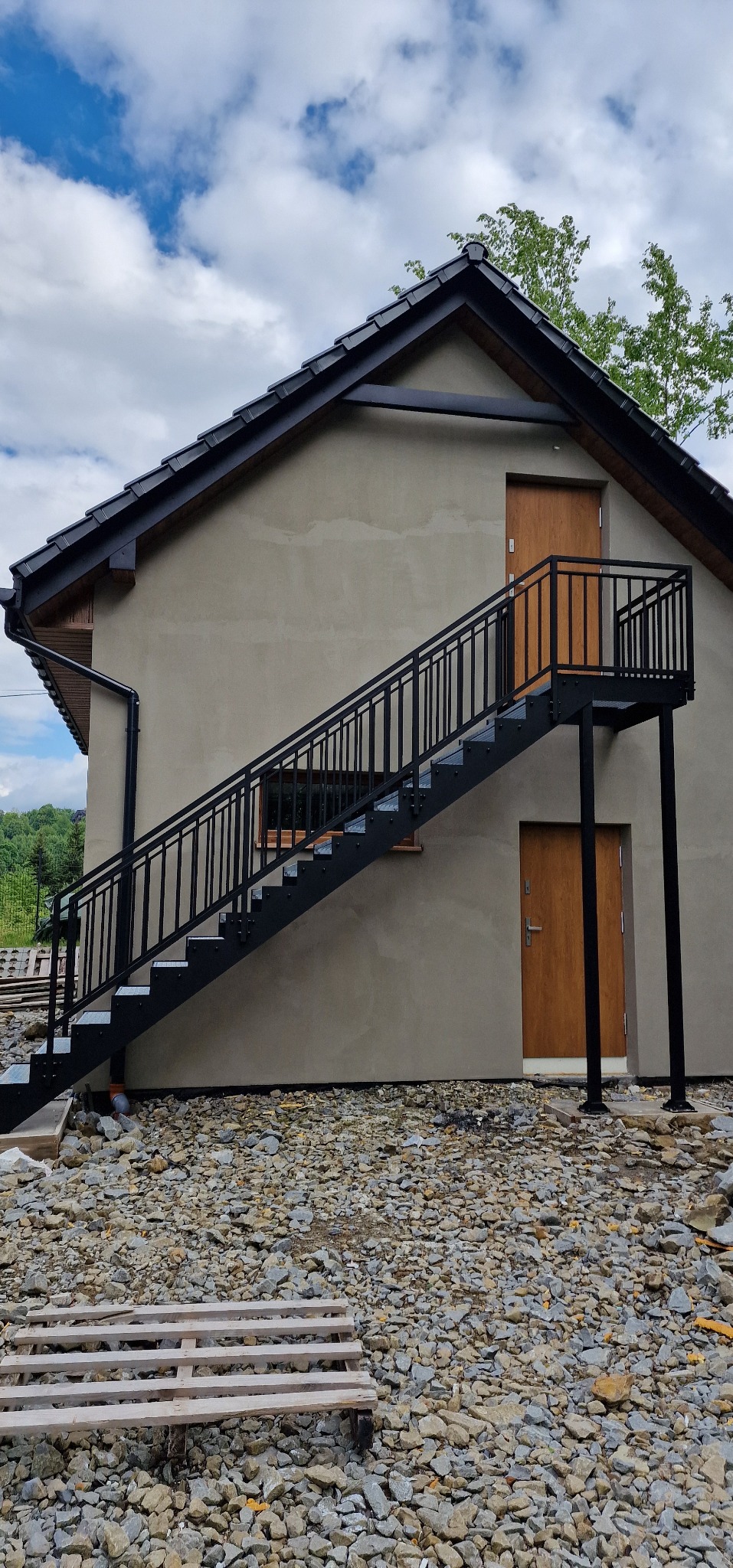 Zewnętrzne schody metalowe z czarną balustradą prowadzące do drzwi na piętrze budynku z szarą elewacją. Widoczne kamienie na podłożu i fragmenty zieleni w tle.