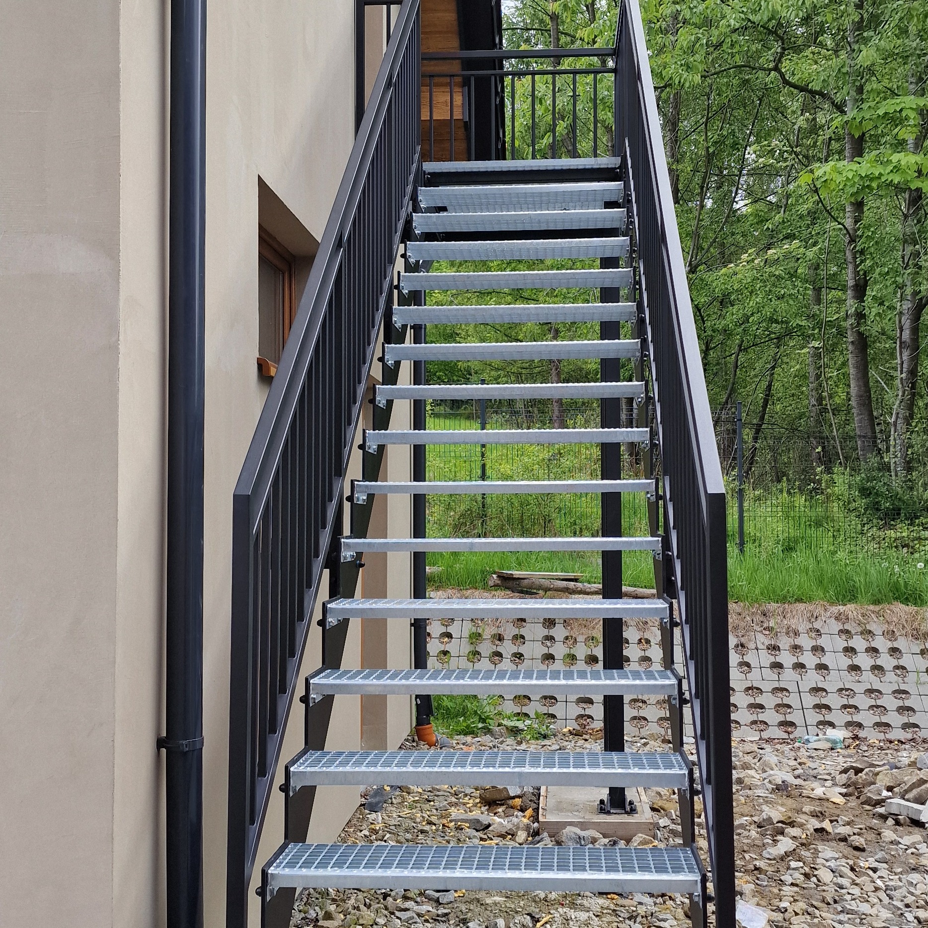 Zewnętrzne schody metalowe z czarną poręczą, prowadzące na piętro budynku w Budzowie. Widok z dołu, na tle zieleni.