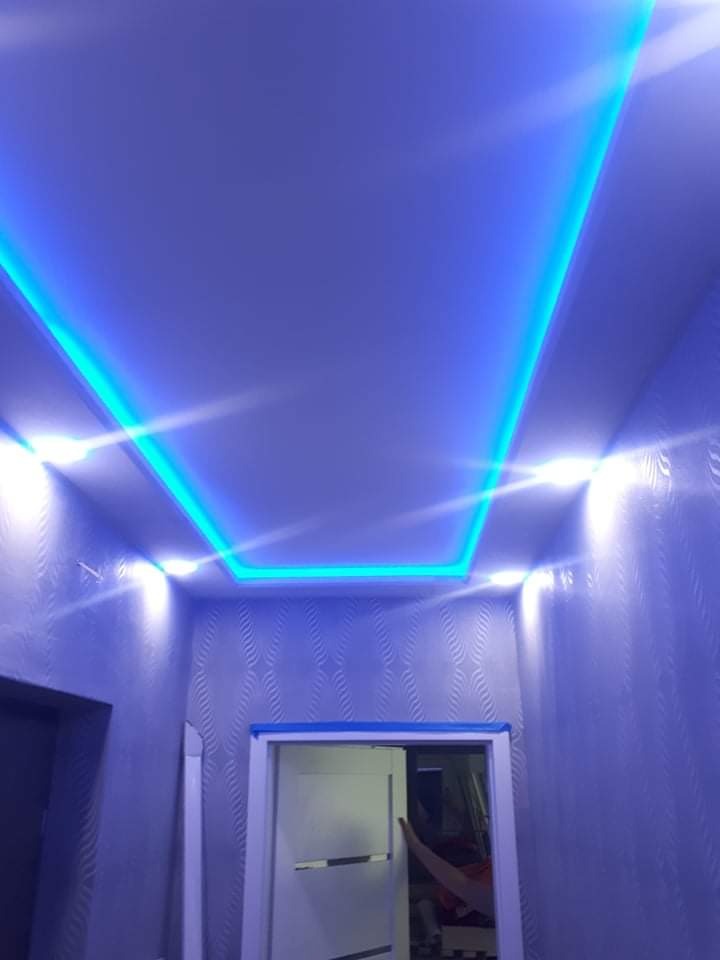 Pokój z niebieskim oświetleniem LED na suficie, wzorzysta tapeta na ścianach, widoczny fragment drzwi z ościeżnicą.