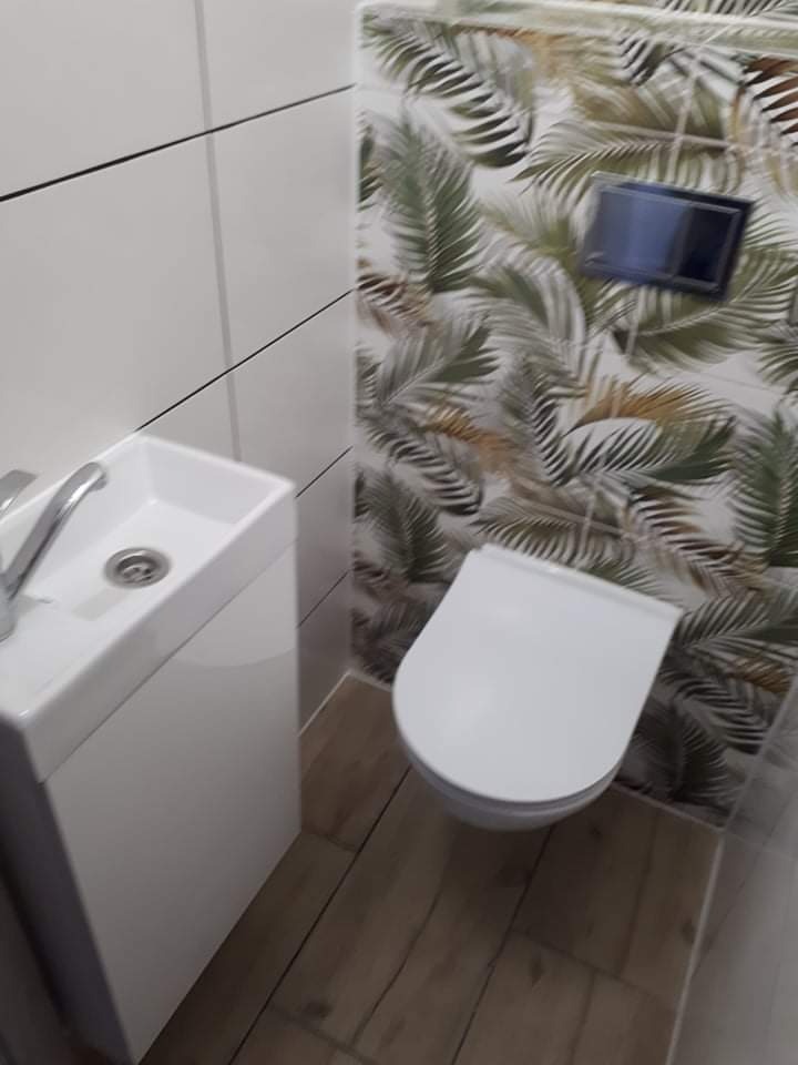 Nowoczesna łazienka z białą ceramiką, umywalką nablatową i toaletą podwieszaną, wykończona płytkami imitującymi drewno na podłodze i tropikalnym wzorem na ścianie za toaletą.