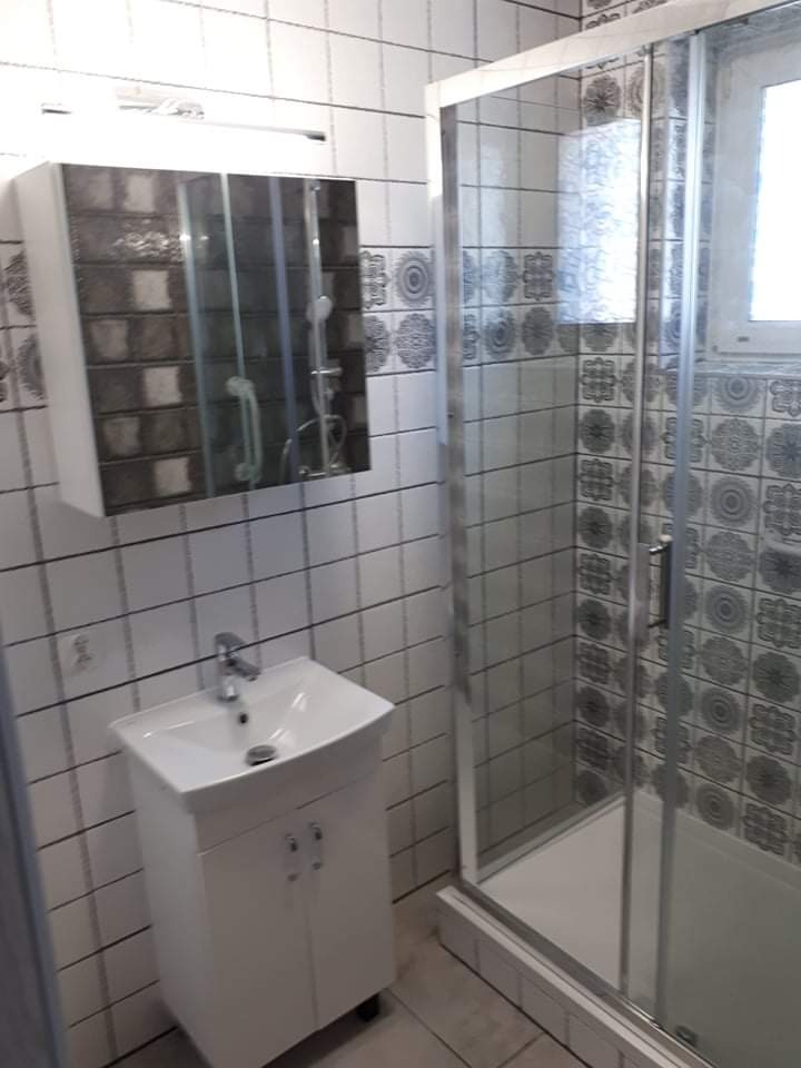 Umywalka z szafką i lustro z oświetleniem nad nią, kabina prysznicowa w łazience wykończonej białymi płytkami z szarym wzorem.