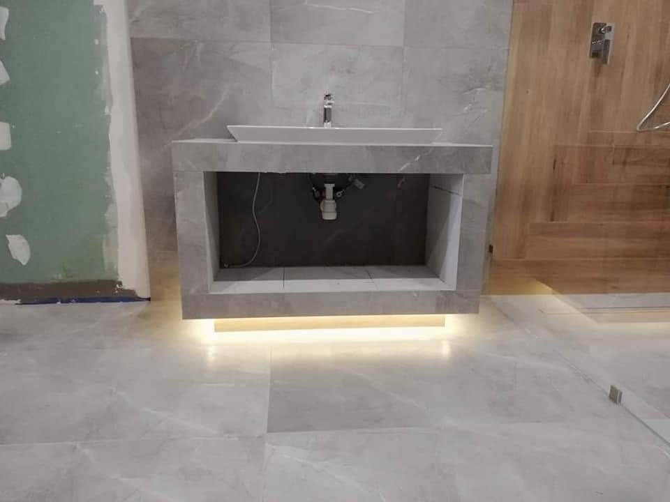 Łazienka w trakcie remontu: umywalka nablatowa z szarą obudową z płytek, podświetlenie LED pod obudową, szare płytki na podłodze i ścianie, fragment ściany w trakcie przygotowania do wykończenia.