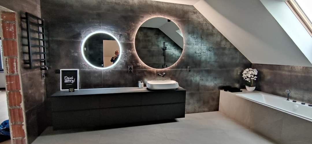 Nowoczesna łazienka na poddaszu z grafitowymi płytkami imitującymi metal, okrągłymi lustrami z podświetleniem LED, białą umywalką nablatową i wanną, oraz grzejnikiem drabinkowym na tle surowej...