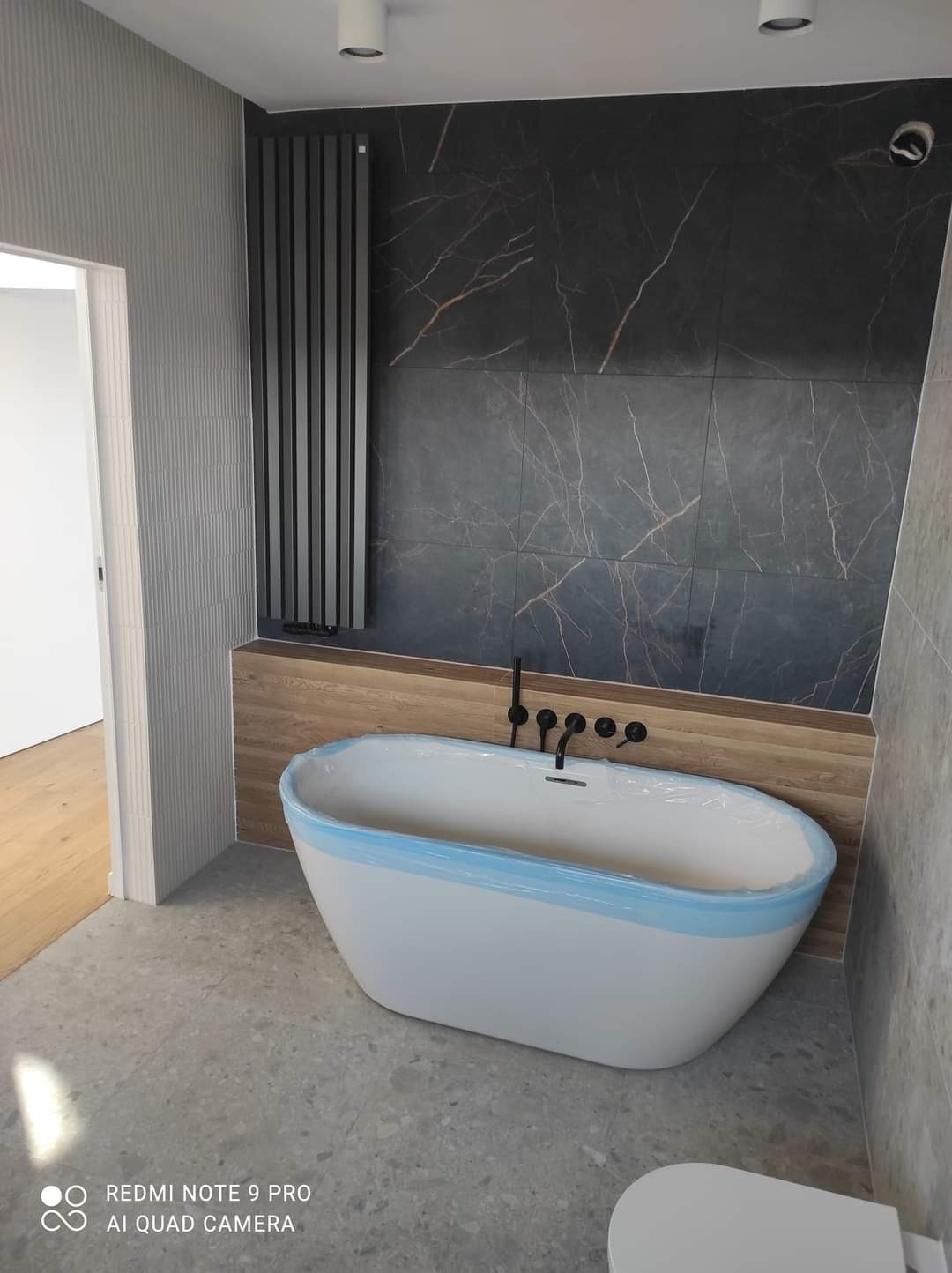 Nowoczesna łazienka z wolnostojącą białą wanną zabezpieczoną folią, czarną armaturą, grzejnikiem panelowym i marmurowymi płytkami na ścianie.