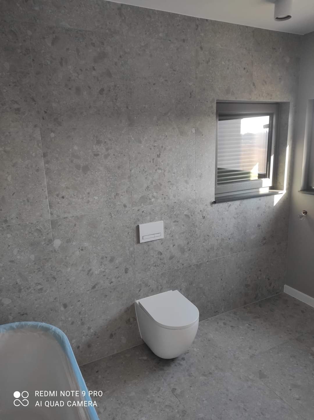 Wykończona łazienka z szarymi płytkami imitującymi lastryko na ścianach i podłodze, wisząca toaleta z białym siedziskiem i spłuczką pod oknem z żaluzjami.