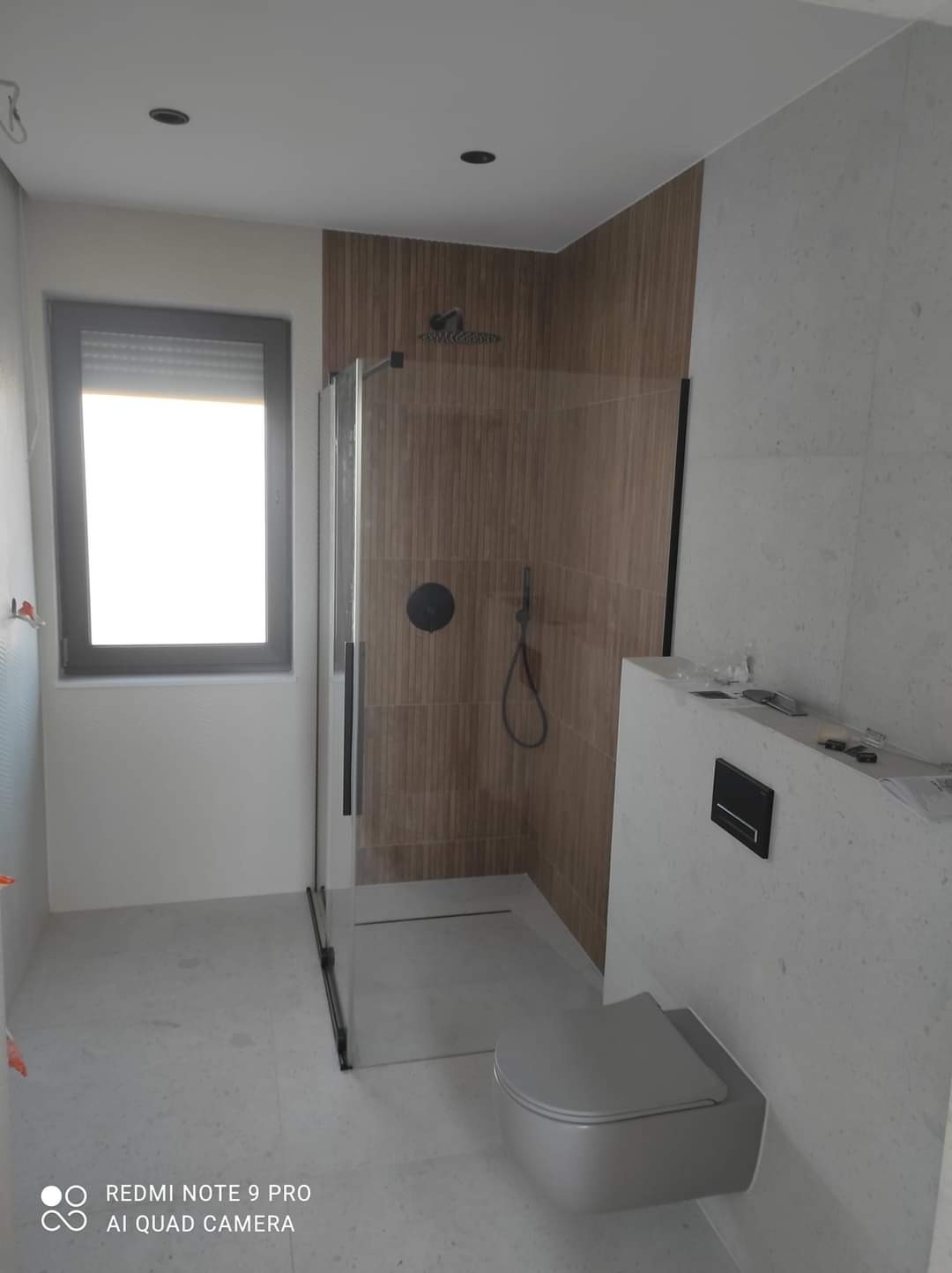 Nowoczesna łazienka z kabiną prysznicową typu walk-in z czarnymi ramami, brązowe płytki imitujące drewno pod prysznicem, wisząca toaleta i jasne płytki podłogowe, okno z roletami.