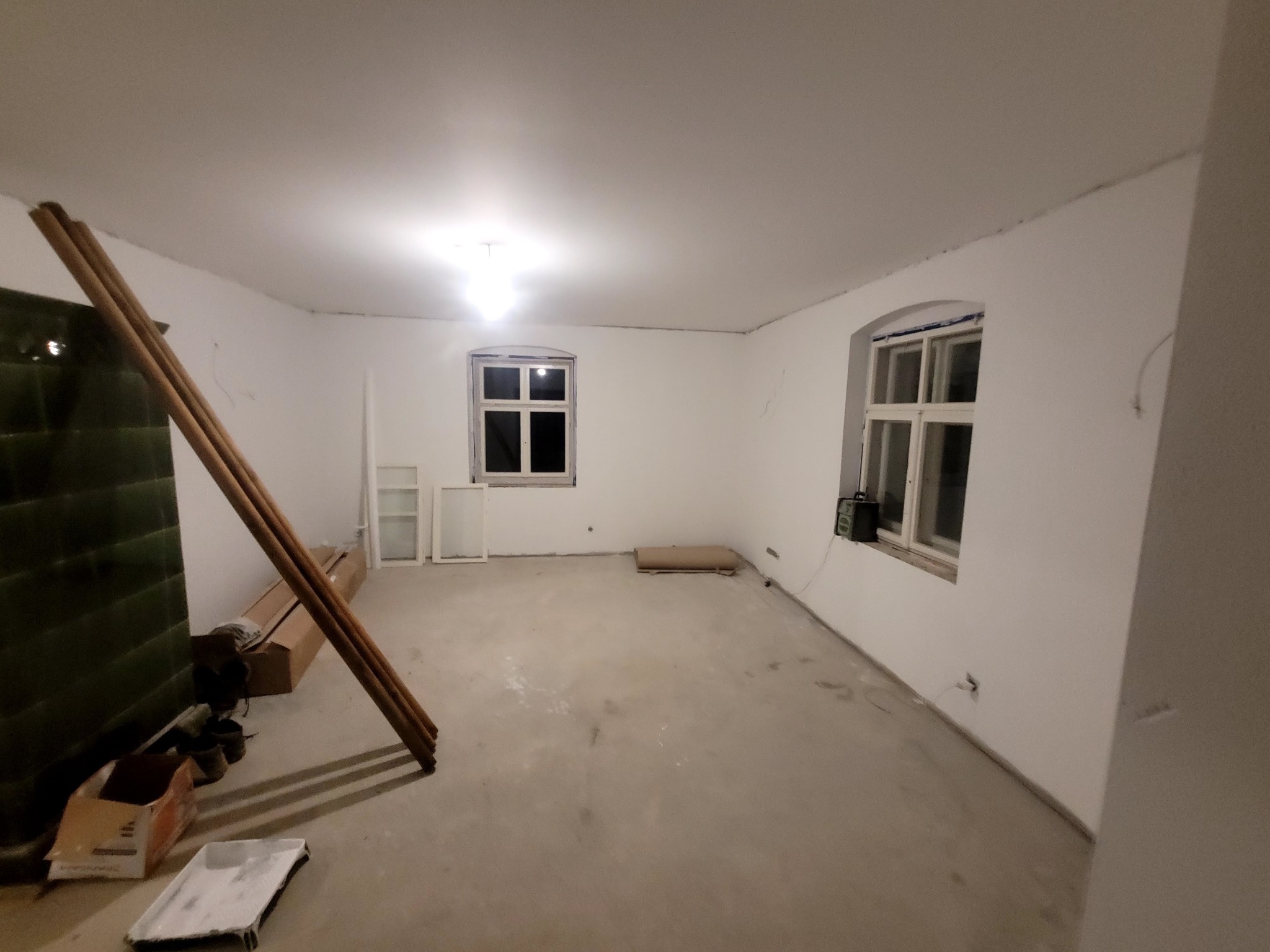 Pusty pokój w trakcie prac przygotowawczych: surowe ściany, zaklejone okna, widoczne narzędzia malarskie i listwy przypodłogowe, przygotowany do dalszych prac wykończeniowych.