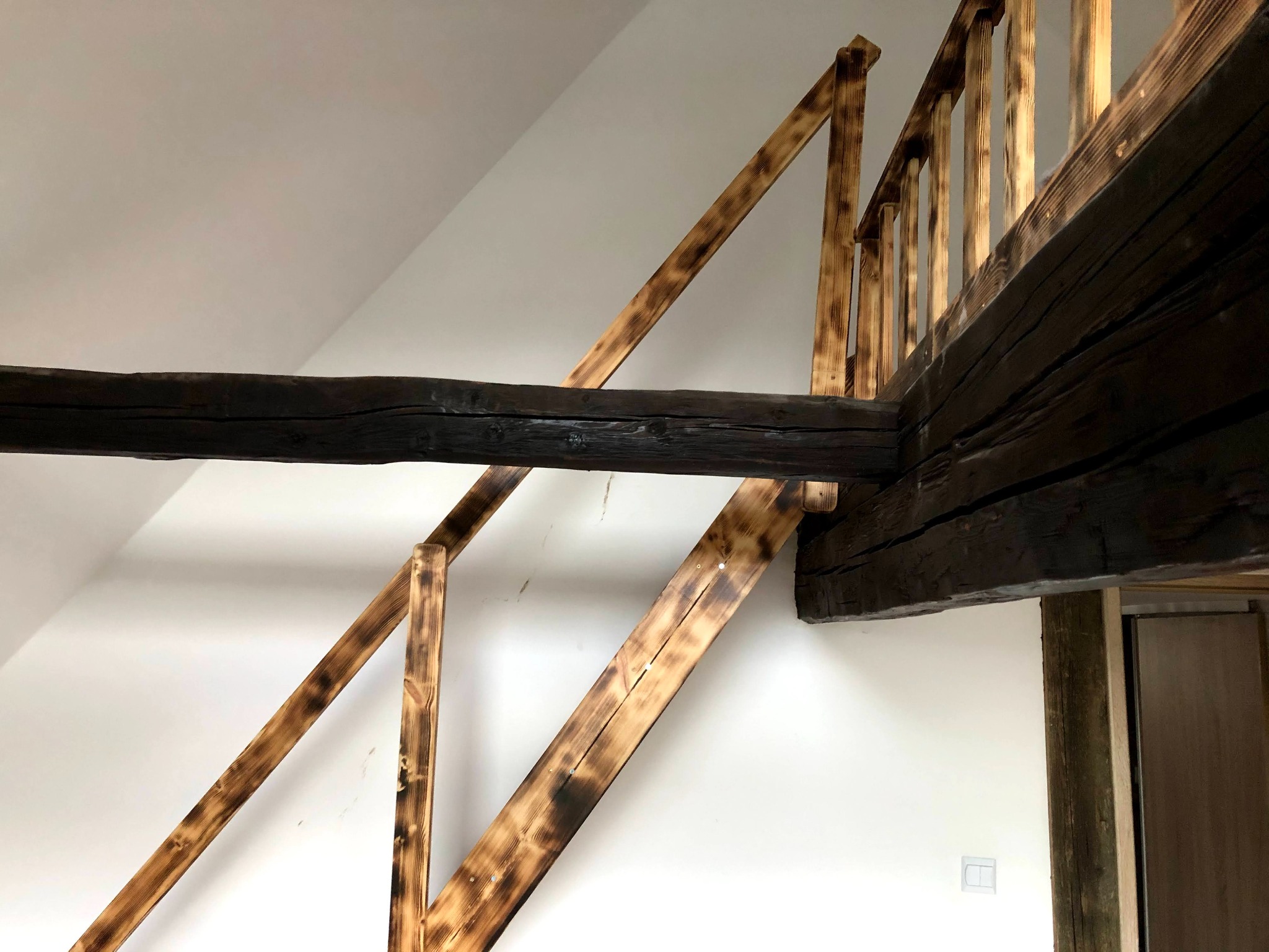 Drewniane elementy konstrukcyjne poddasza z widocznymi śladami opalania, kontrastujące z białą ścianą i fragmentem drewnianej balustrady.