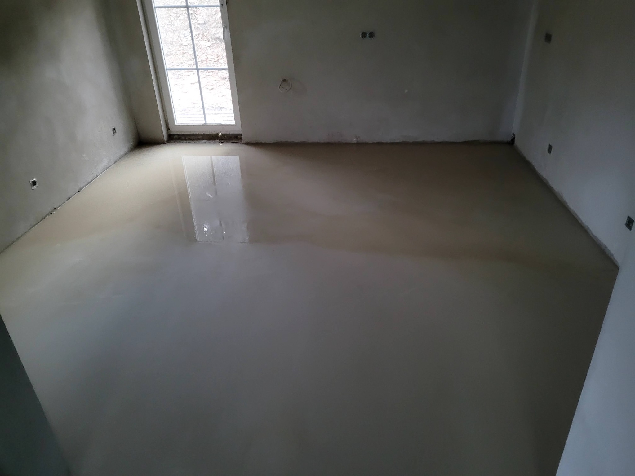 Wylewka samopoziomująca na podłodze w pustym, surowym pomieszczeniu z oknem, ściany przygotowane do dalszych prac wykończeniowych.