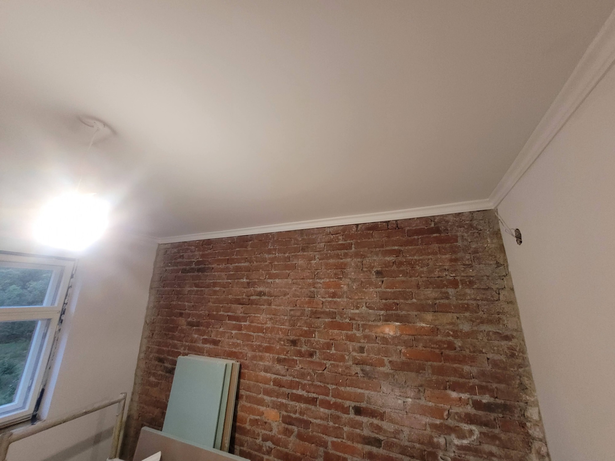 Fragment pokoju z odsłoniętą czerwoną cegłą na ścianie, częściowo otynkowaną, z widocznymi płytami izolacyjnymi i listwą dekoracyjną pod sufitem.