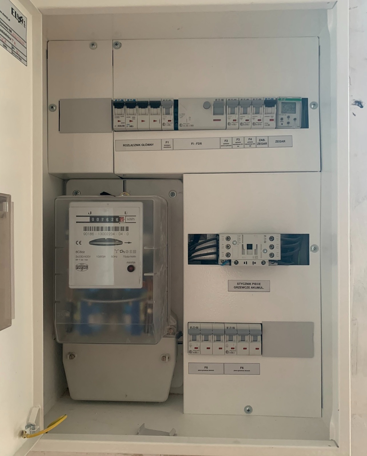 Biała skrzynka elektryczna z licznikiem i bezpiecznikami, widoczne oznaczenia: rozłącznik główny, stycznik pieca grzewczego akumulacyjnego.