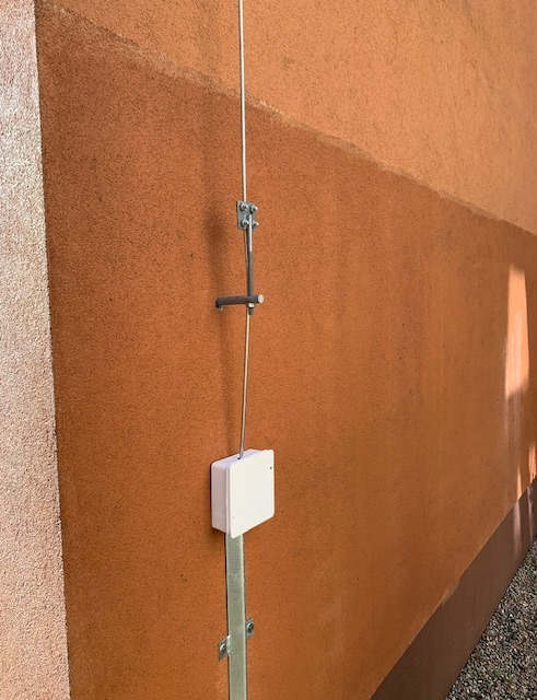 Zewnętrzna instalacja odgromowa z białą puszką i metalowym wspornikiem na pomarańczowej fasadzie budynku, widoczna z dołu.