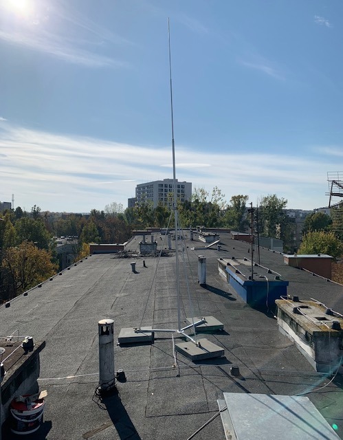 Instalacja odgromowa na dachu budynku, widok z góry. Metalowa konstrukcja z masztem na tle błękitnego nieba i zabudowy miejskiej. Widoczne elementy mocujące i kable.