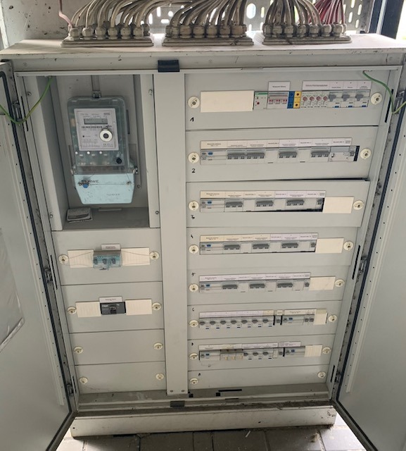 Otwarta skrzynka elektryczna z licznikiem i bezpiecznikami, widoczne kable w górnej części. Szare elementy, białe napisy na bezpiecznikach.