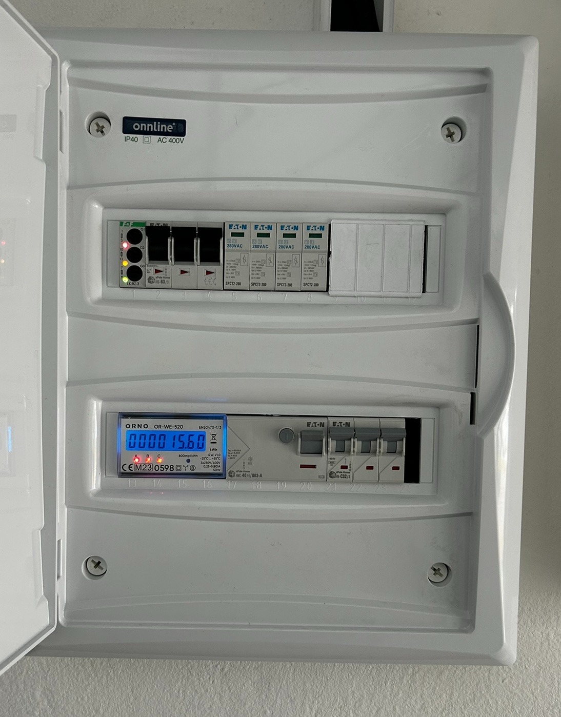 Otwarta, biała skrzynka elektryczna z zainstalowanymi bezpiecznikami Eaton i licznikiem Orno OR-WE-520, wskazującym zużycie energii. Widoczne diody i oznaczenia na panelu.