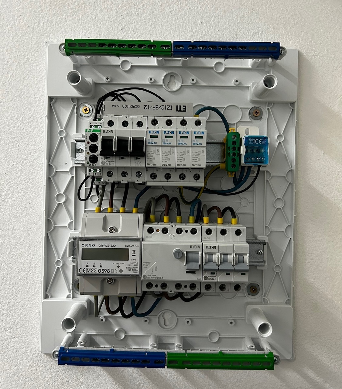 Rozdzielnica elektryczna z zamontowanymi bezpiecznikami, licznikiem i przewodami. Widoczne elementy marki Eaton i Orno. Biała obudowa na jasnym tle ściany.