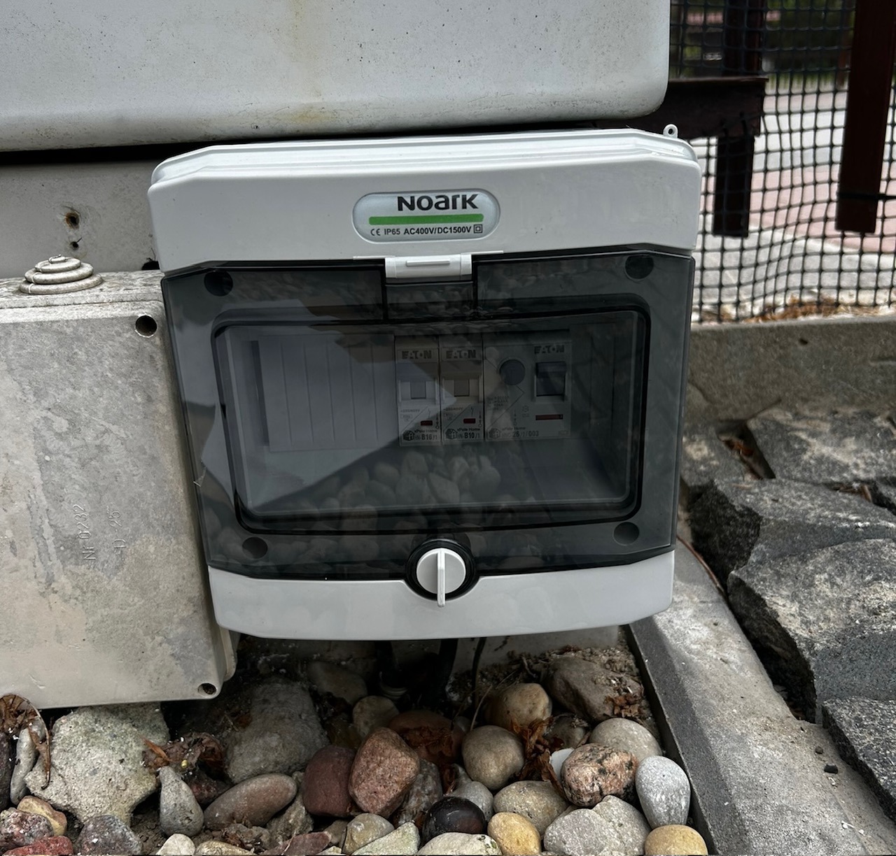 Szara skrzynka elektryczna Noark z przezroczystą osłoną, zamontowana na zewnątrz, widoczne bezpieczniki Eaton, ułożona na kamieniach. Instalacja z widoczną tabliczką znamionową.
