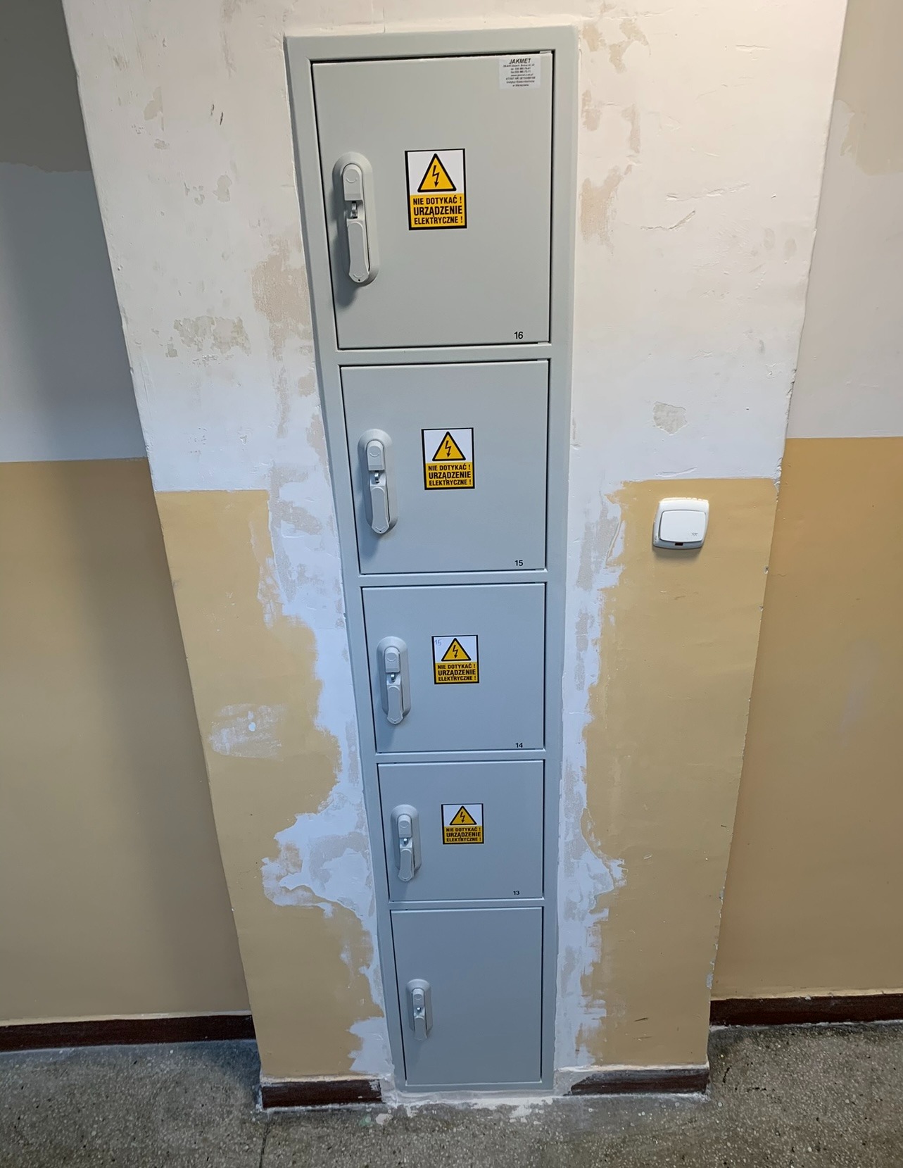 Szara, czterosegmentowa skrzynka elektryczna z ostrzeżeniami 'Nie dotykać! Urządzenie elektryczne!' na ścianie w trakcie malowania, widoczny fragment włącznika.