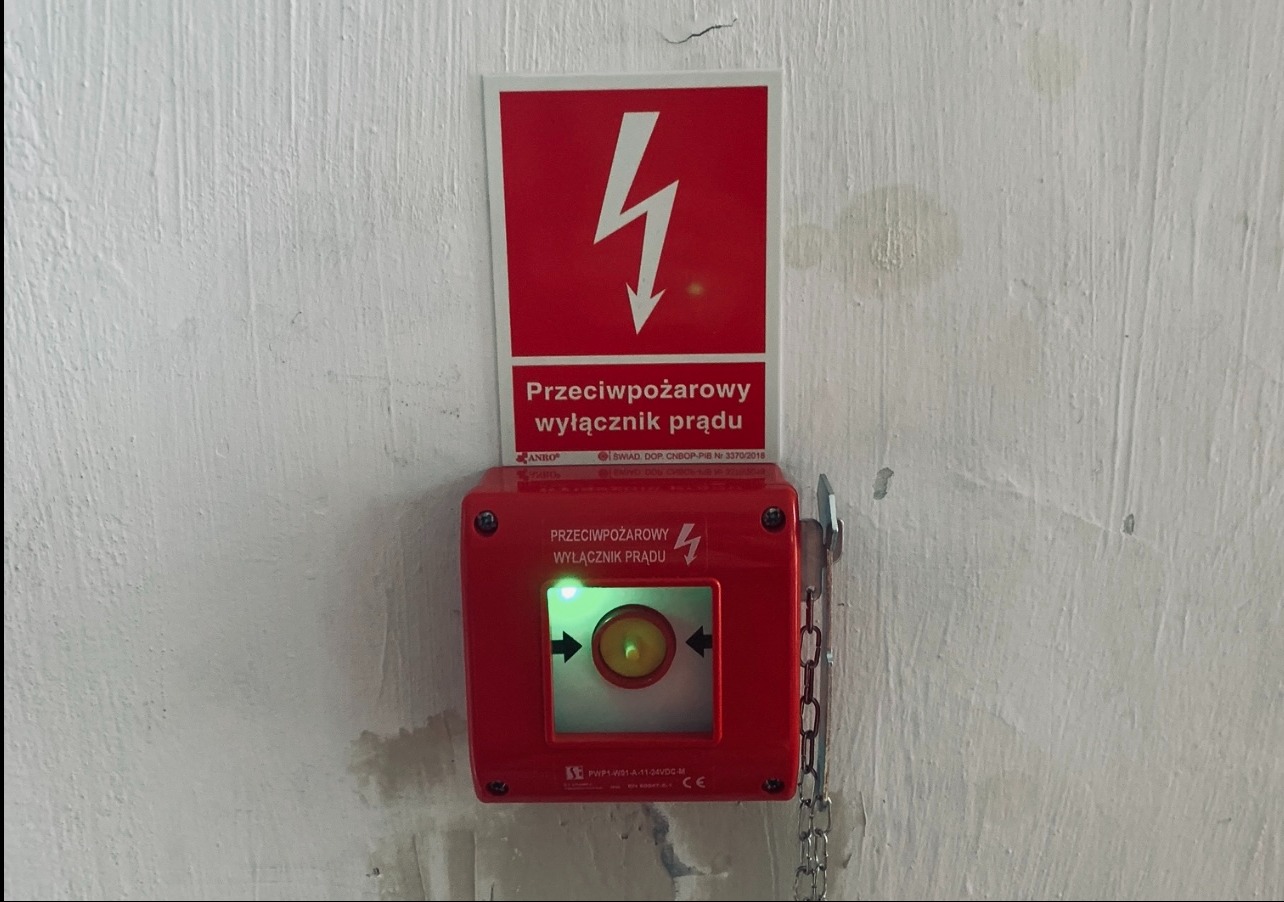 Czerwony, naścienny, przeciwpożarowy wyłącznik prądu z zieloną diodą, pod znakiem ostrzegawczym z symbolem błyskawicy. Zamontowany na pomalowanej ścianie.