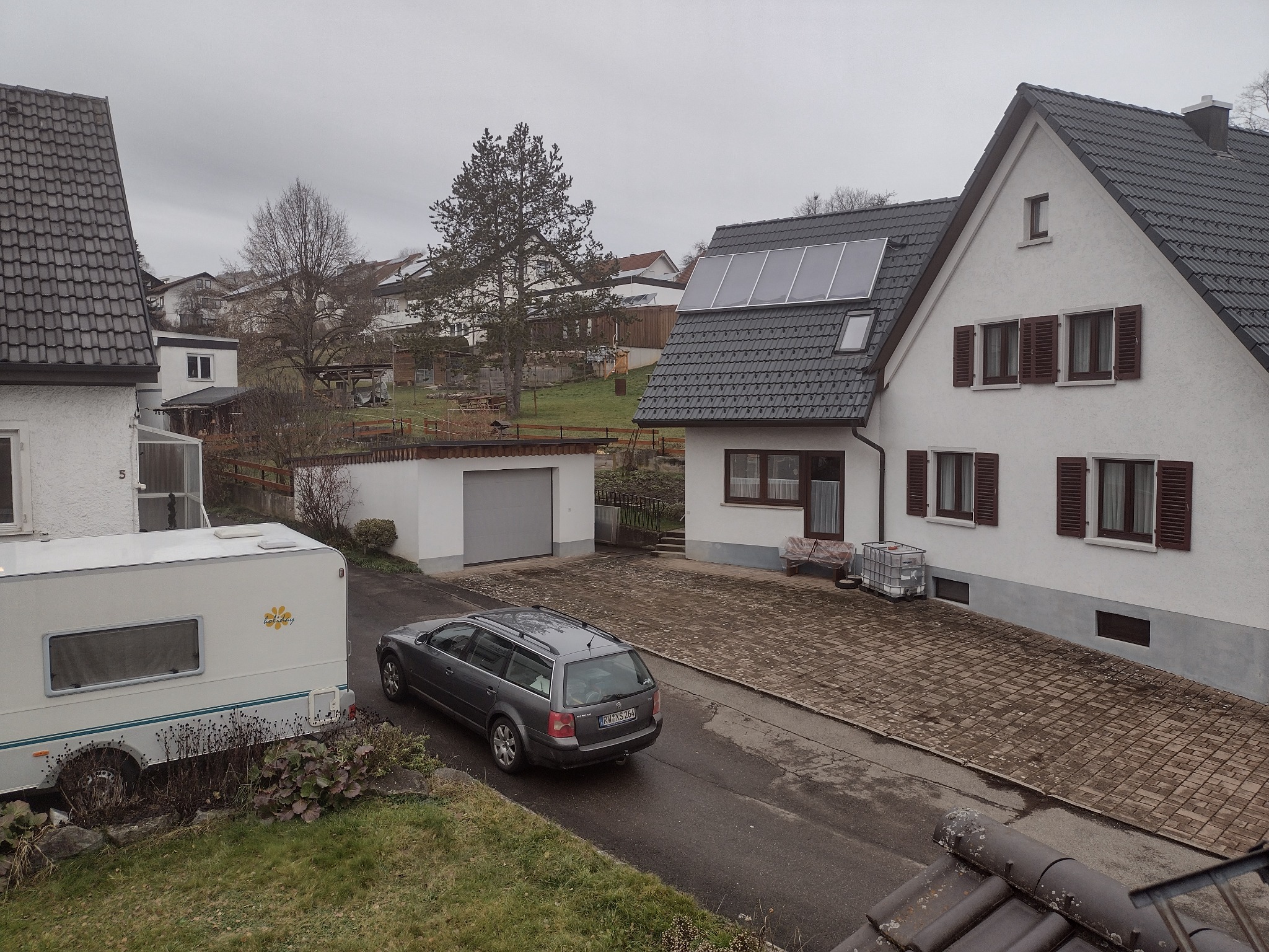 Szary samochód kombi zaparkowany na podjeździe przed domem z panelami słonecznymi na dachu i przyczepą kempingową z logo 'Holiday'.