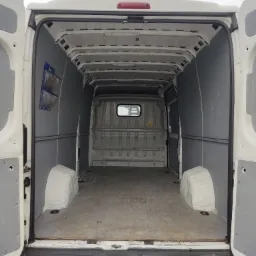 Wnętrze pustego, białego furgonetki z otwartymi tylnymi drzwiami, widoczne szare panele ochronne i uchwyty na ścianach.