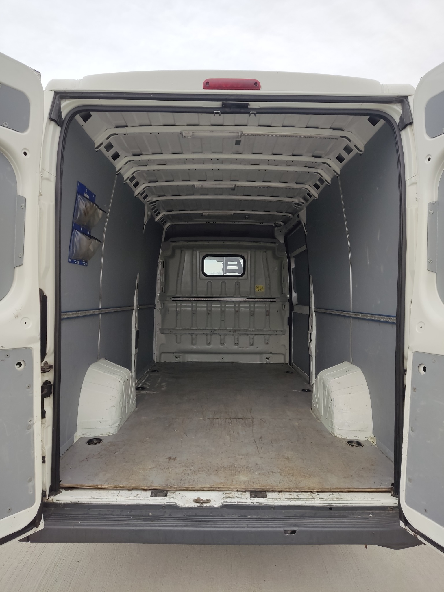 Wnętrze pustego, białego furgonetki z otwartymi tylnymi drzwiami, widoczne szare panele ochronne i uchwyty na ścianach.