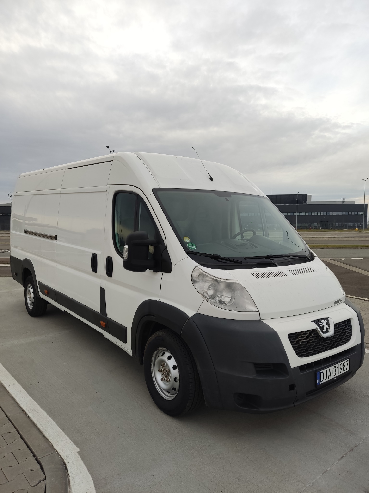 Biały Peugeot Boxer na parkingu lotniskowym, widok z przodu, pochmurne niebo