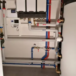 Instalacja z trzema białymi modułami grzewczymi Kolton, zbiornikiem akumulacyjnym i rurami oznaczonymi strzałkami kierunku przepływu, zamontowana w pomieszczeniu z szarą podłogą.