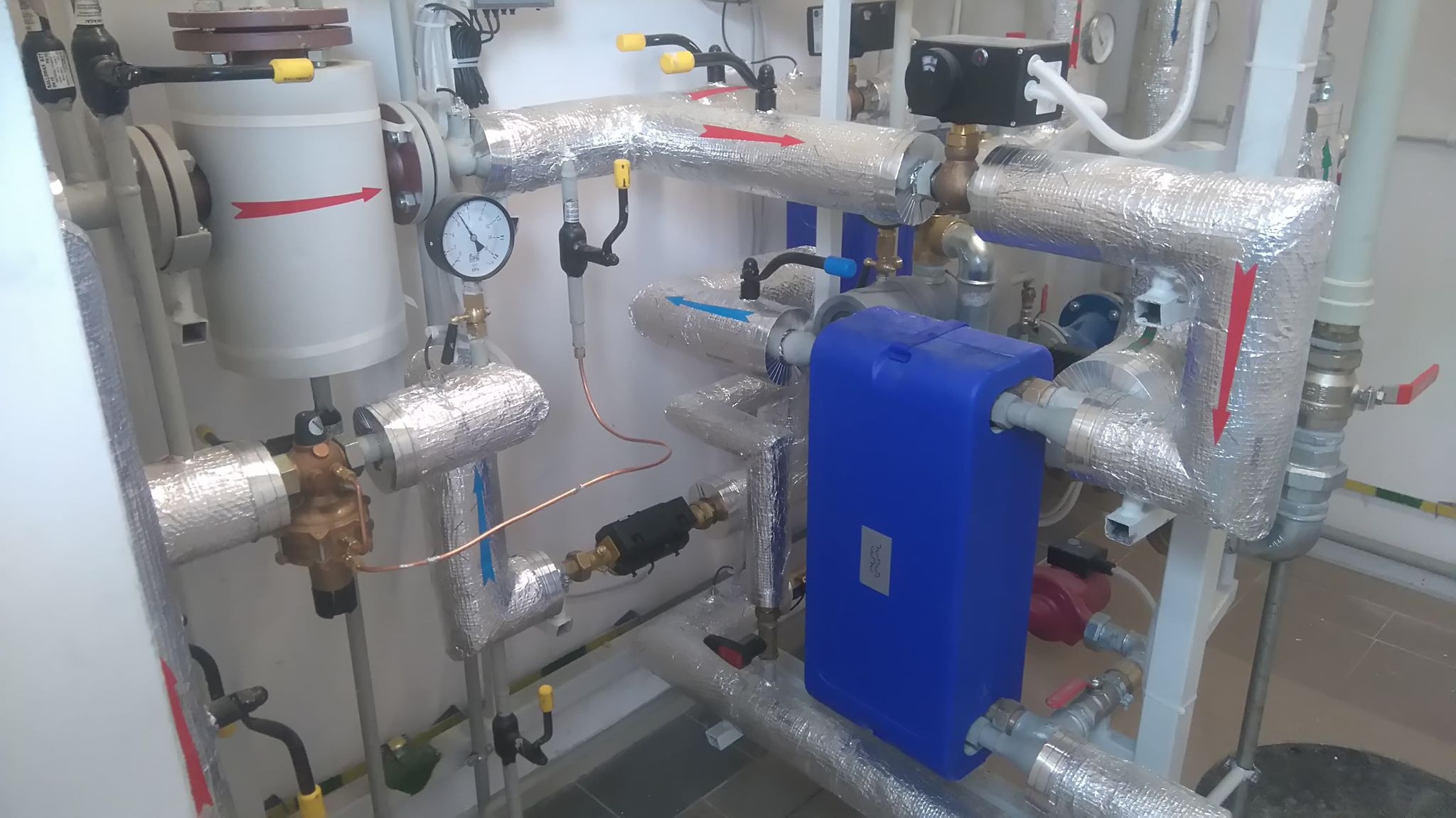 Instalacja centralnego ogrzewania z rurami w srebrnej izolacji, zbiornikiem wyrównawczym i manometrem, widoczne strzałki wskazujące kierunek przepływu.