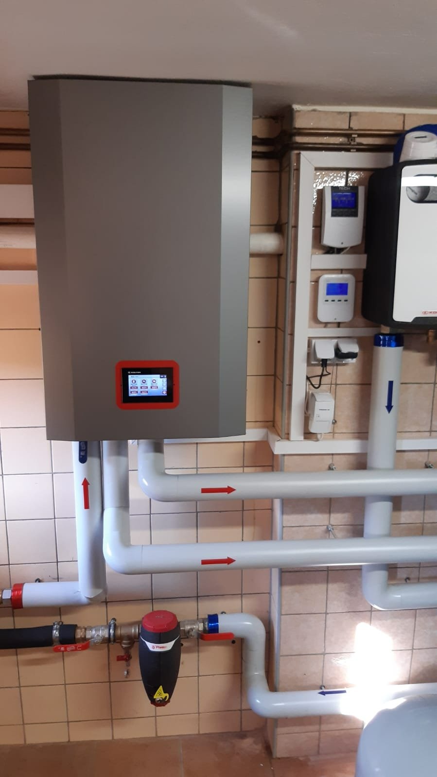 Szara jednostka grzewcza z czerwonym panelem sterowania, rury instalacyjne z czerwonymi strzałkami wskazującymi kierunek przepływu, filtr na rurze oraz dodatkowe urządzenia sterujące na ścianie...