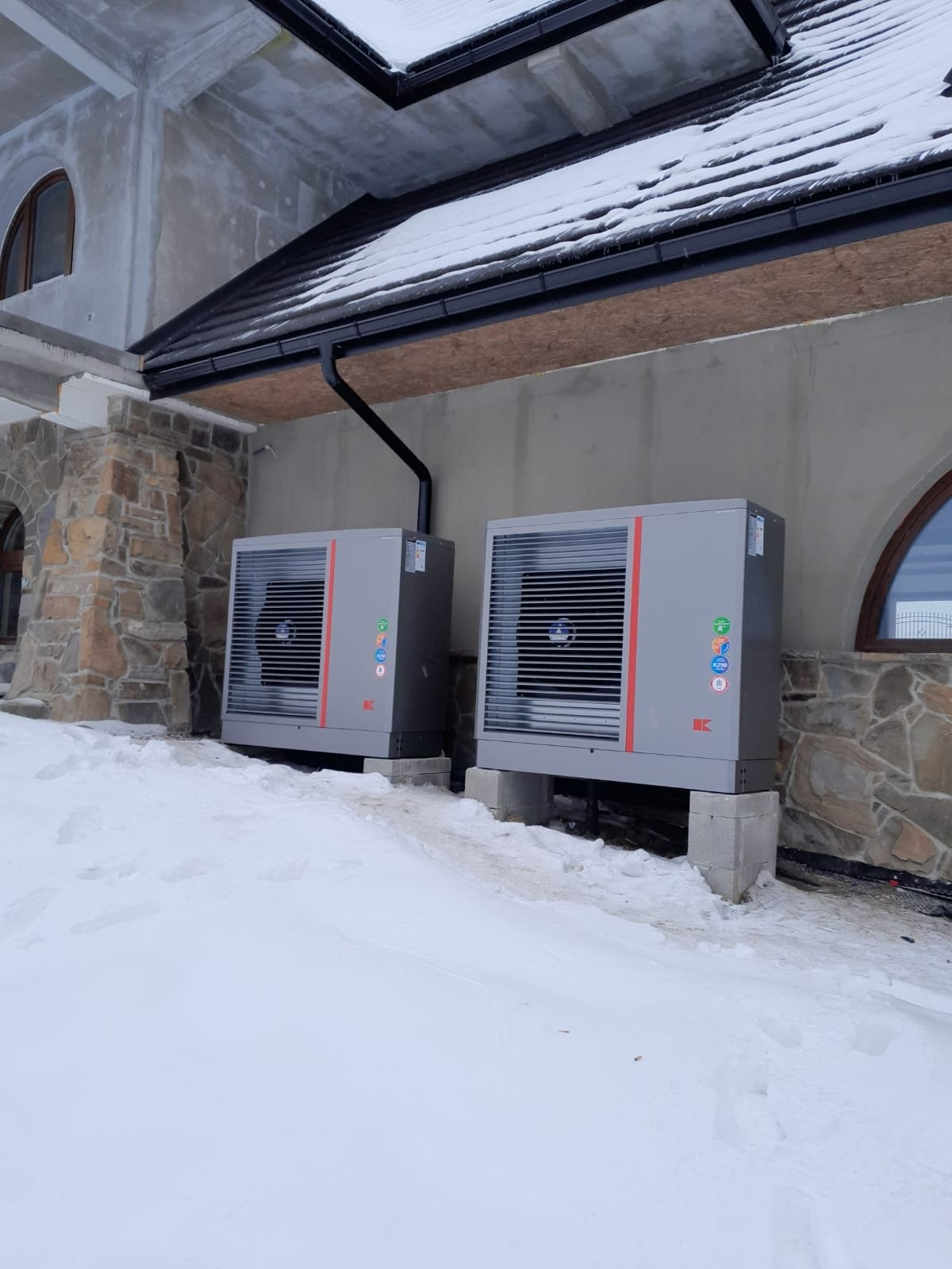 Dwie szare jednostki zewnętrzne z czerwonym paskiem, zamontowane na betonowych podstawach przy budynku w zimowym krajobrazie, widoczne etykiety i logo producenta.