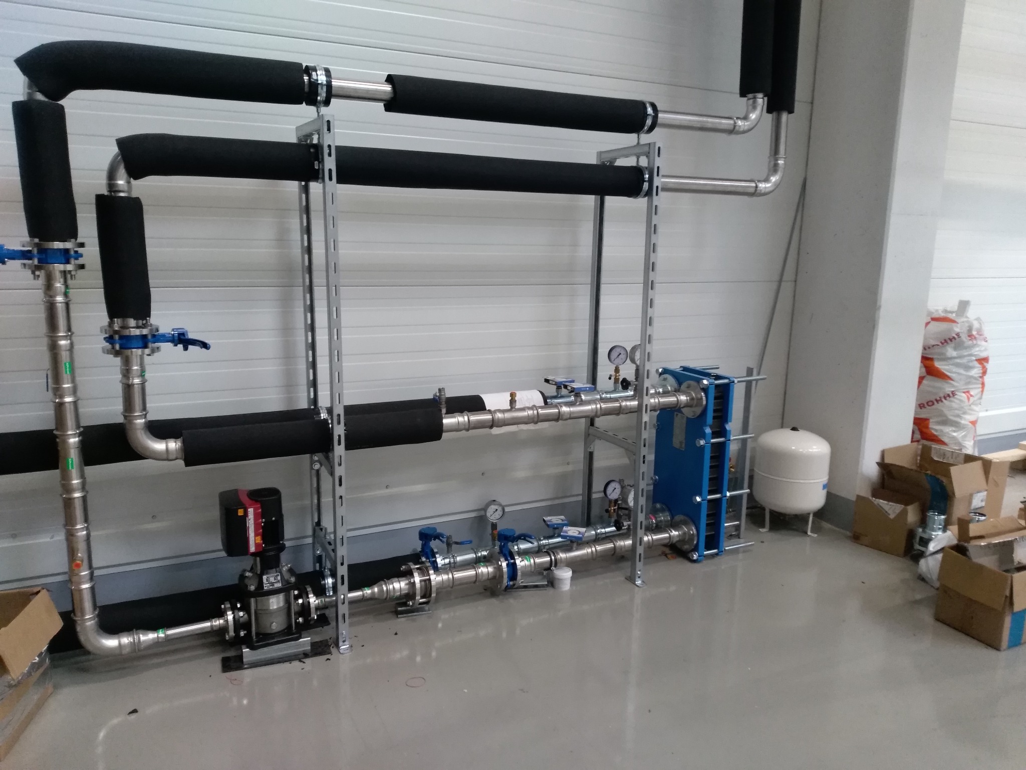 Złożona instalacja rur, zaworów i urządzeń grzewczych, w tym czarna pompa obiegowa, niebieski wymiennik ciepła płytowy i biały zbiornik wyrównawczy, zamontowana w pomieszczeniu o ścianach z blachy...