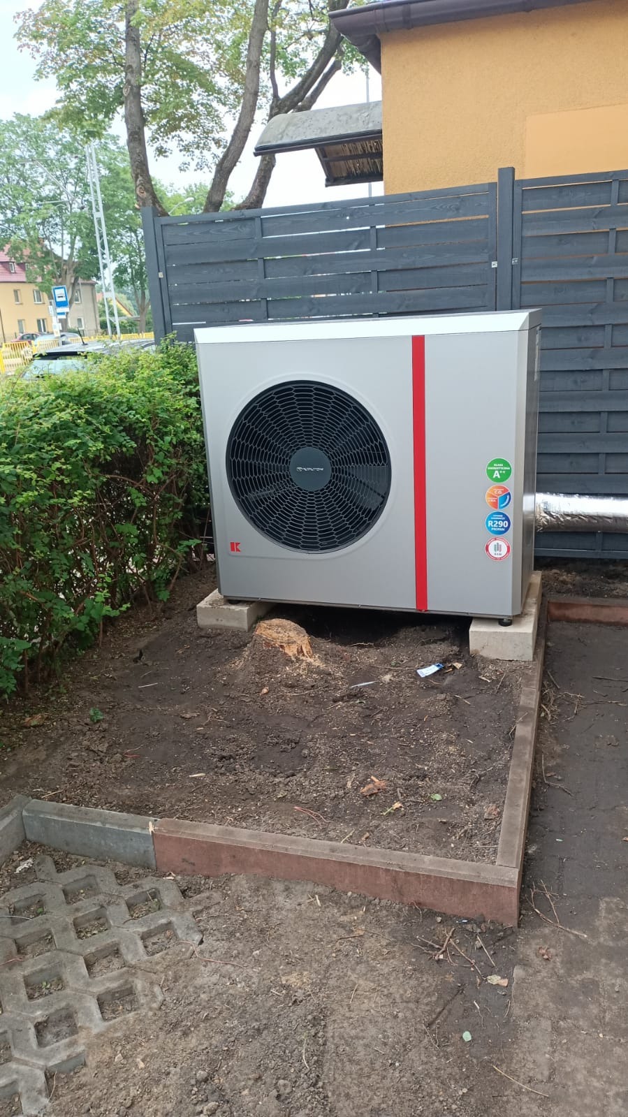 Szara pompa ciepła z czerwoną linią, umieszczona na betonowych podstawach w ogrodzie, widoczna z boku, z szarą rurą wentylacyjną i etykietami energetycznymi.