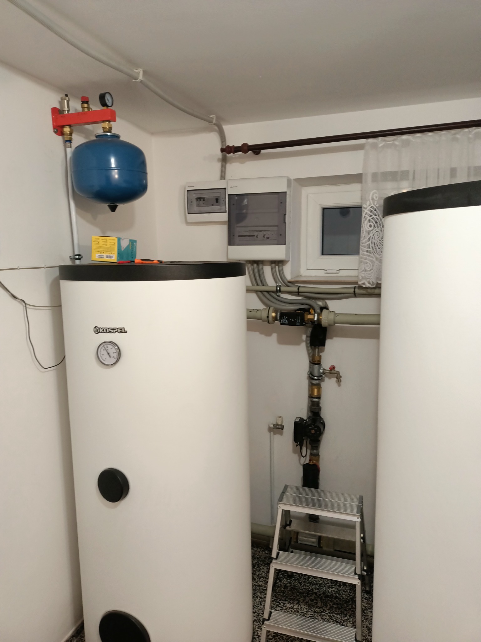 Dwa białe zasobniki ciepłej wody Kospel w kotłowni, z widocznymi elementami instalacji hydrauliczej, niebieskim naczyniem wzbiorczym i drabinką.
