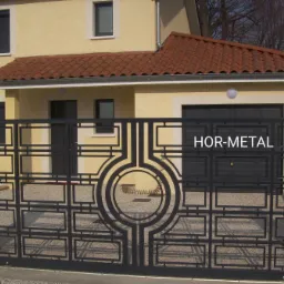 Nowoczesna, czarna brama metalowa z geometrycznym wzorem na tle beżowego domu z czerwoną dachówką.