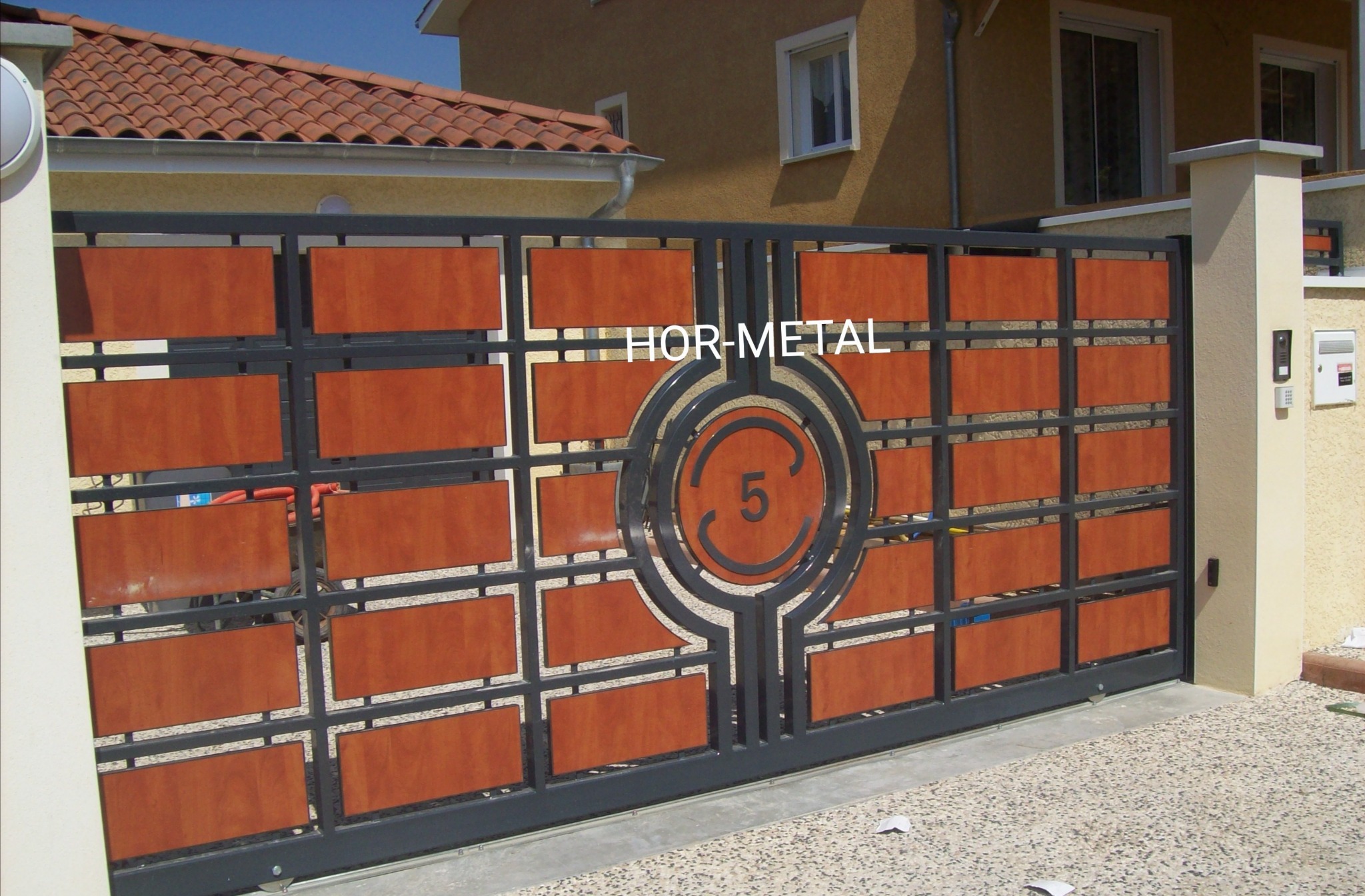 Przesuwna brama wjazdowa z metalową ramą i drewnianymi panelami, z centralnym okrągłym elementem dekoracyjnym z numerem '5', na tle domu z czerwoną dachówką.