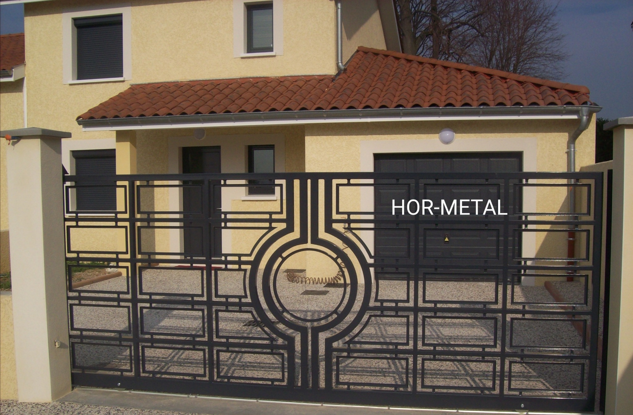 Nowoczesna, czarna brama metalowa z geometrycznym wzorem na tle beżowego domu z czerwoną dachówką.