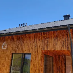 Nowoczesny dom z drewnianą elewacją i instalacją odgromową na dachu, widoczny z niskiej perspektywy, na tle błękitnego nieba.