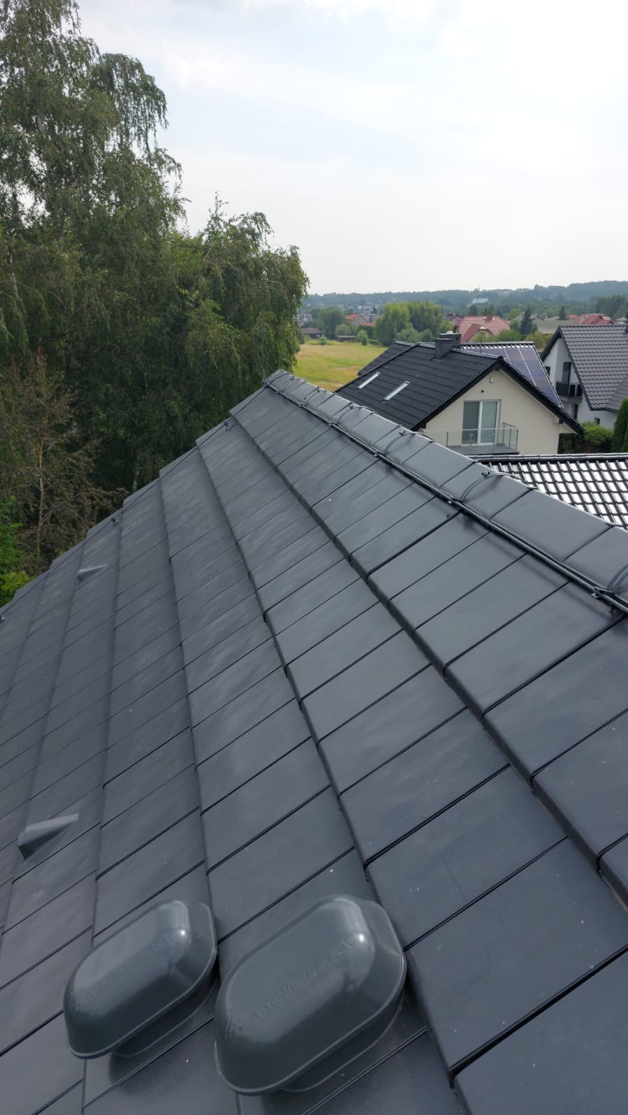 Ciemnoszary dach z dachówki ceramicznej z zamontowanymi elementami instalacji odgromowej, widok z góry na tle zabudowań mieszkalnych i drzew.