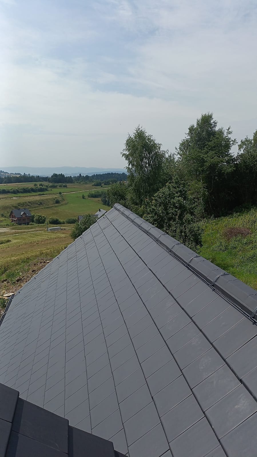 Widok z góry na dach pokryty szarą dachówką, z widocznym systemem zabezpieczenia odgromowego, w tle krajobraz z zabudowaniami i drzewami.