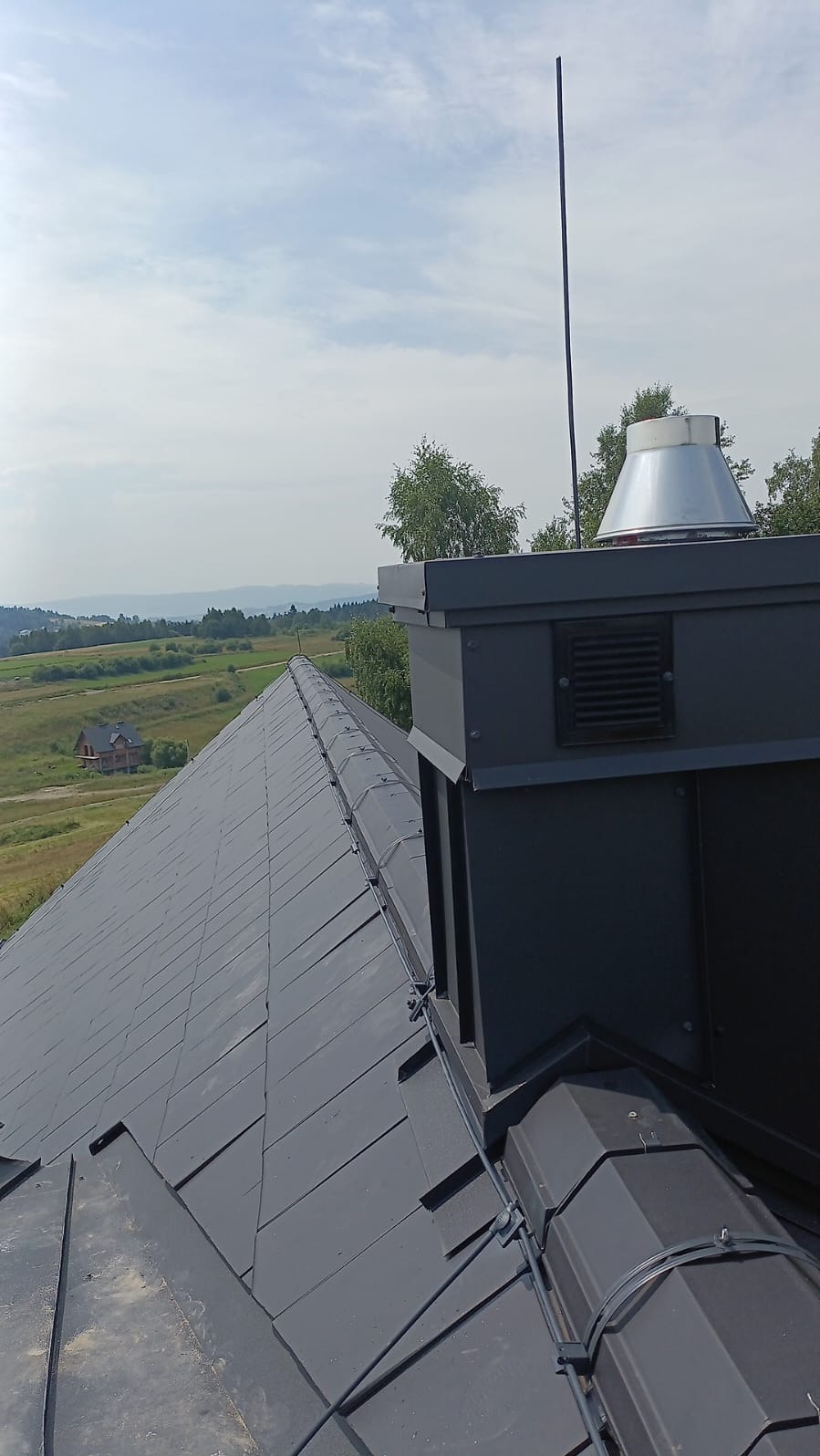 Szary dach z blachy z zamontowanym systemem ochrony przed wyładowaniami atmosferycznymi i kominem na tle pagórkowatego krajobrazu.