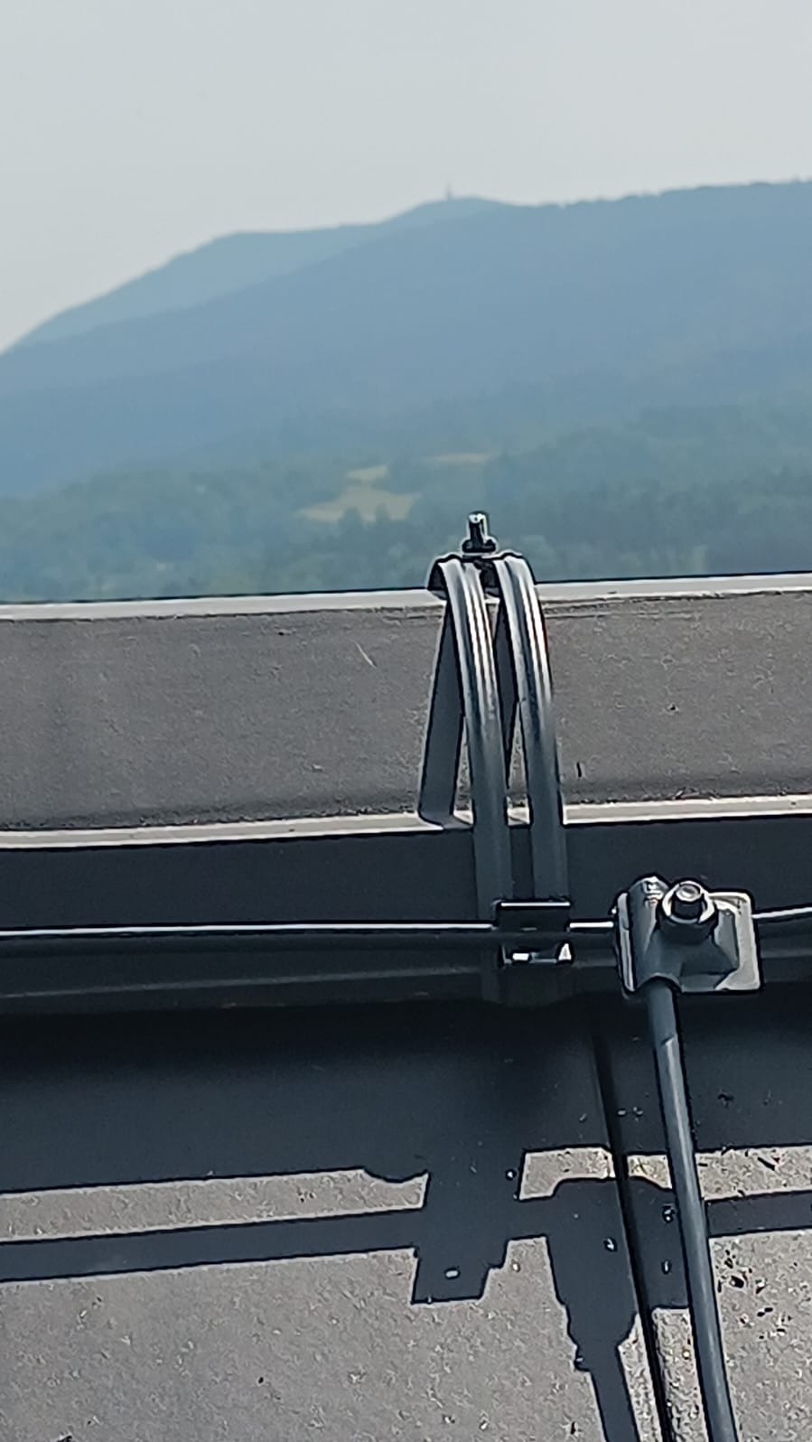 Metalowy uchwyt instalacji odgromowej przymocowany do dachu z widokiem na zalesione wzgórza w oddali.